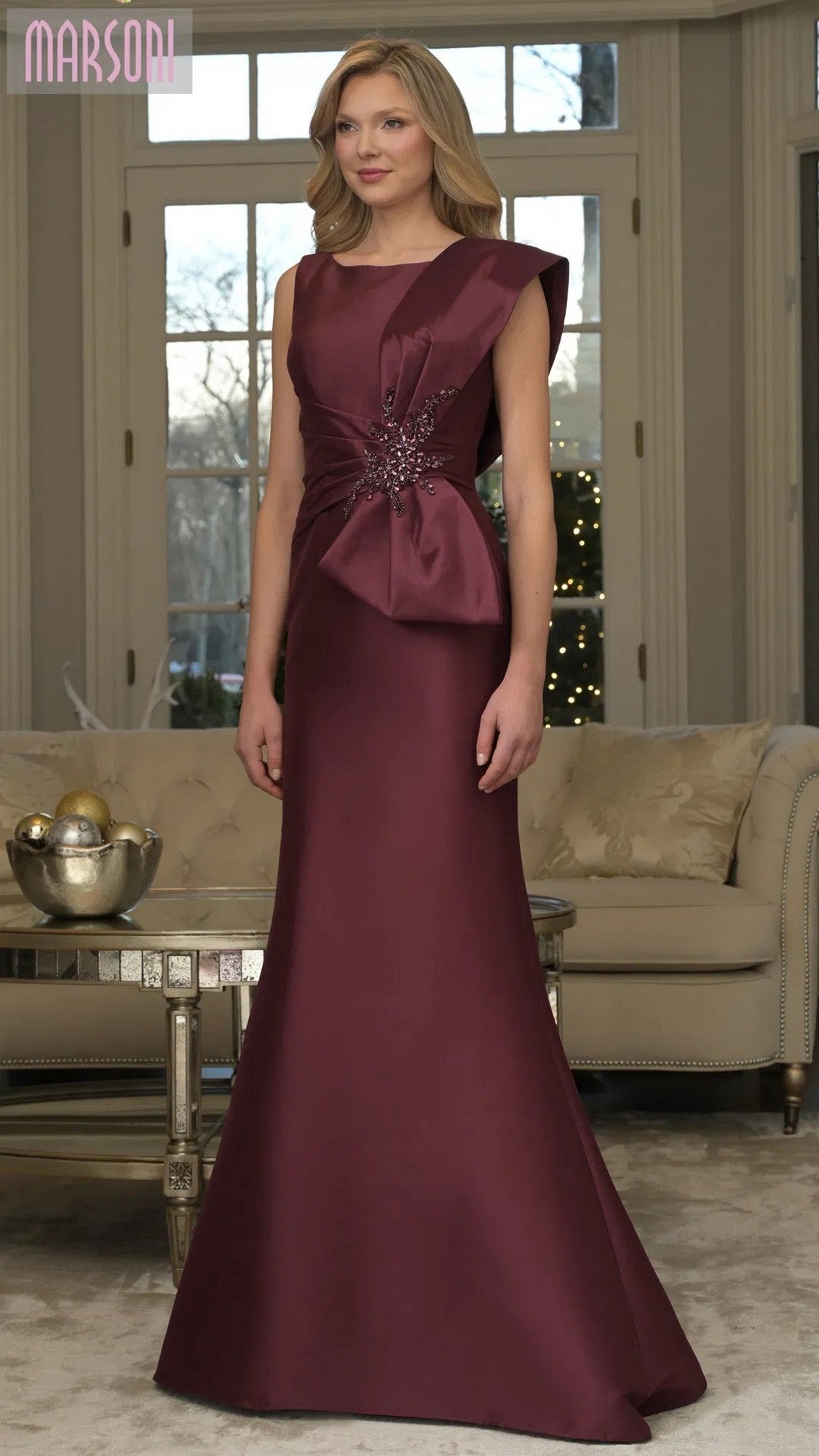 A woman poses by a beige sofa and large windows wearing the Marsoni MV1359 long formal dress in wine, featuring a sleeveless bodice, mermaid skirt, decorative side bow, bead accents, and floor-length silhouette.